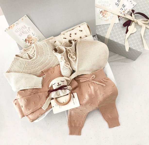 Caja de regalo abierta con ropa de bebé de punto en color salmón y beige, incluyendo patucos, gorro y sonajero de conejo.
